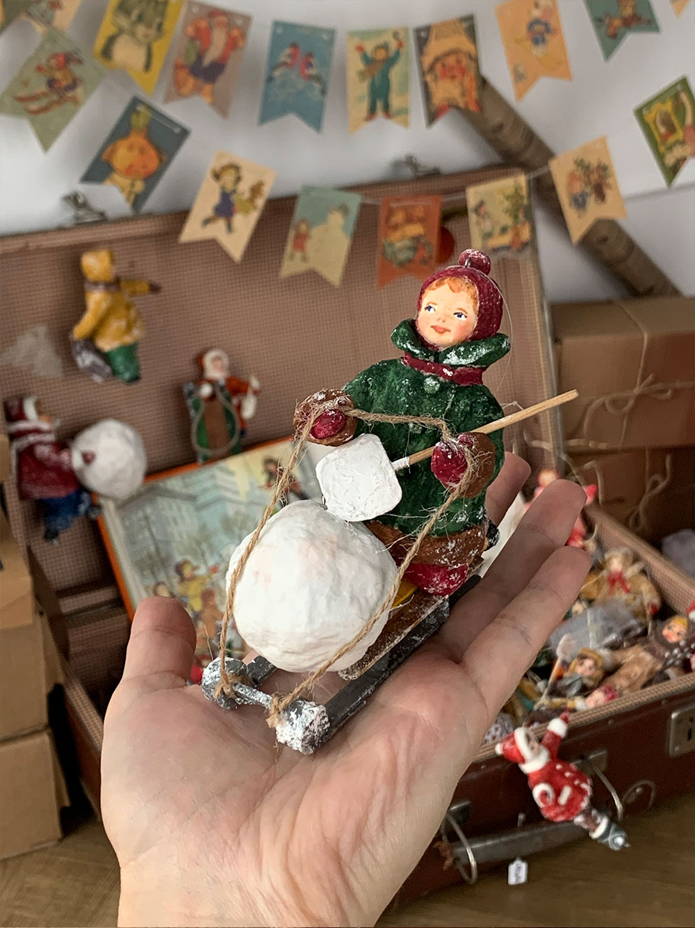 Елочная игрушка в виде ребенка на санках, который везет снежный ком с лопаткой в руке.