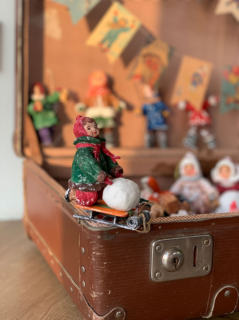 Елочная игрушка в виде девочки с санками на фоне открытого чемодана.