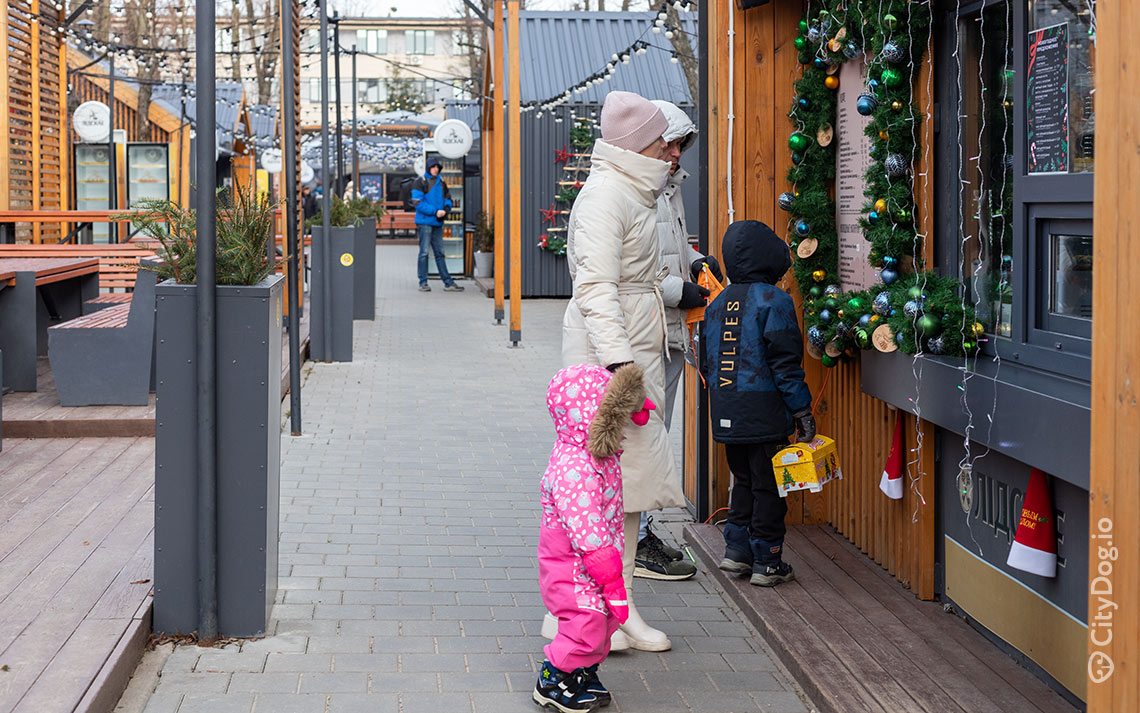 Женщина в белом пальто держит за руку ребенка в розовой куртке, рядом стоит ребенок в синей куртке.