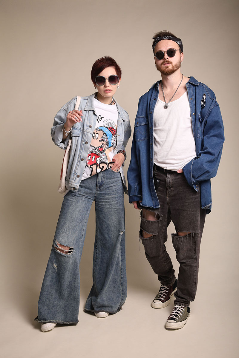 На фото стоят парень и девушка. На девушке – широкие синие джинсы с разрезами и светлая джинсовка. На парне – белая майка, темно-синяя рубашка, и прямые темно-серые джинсы с дырками. 