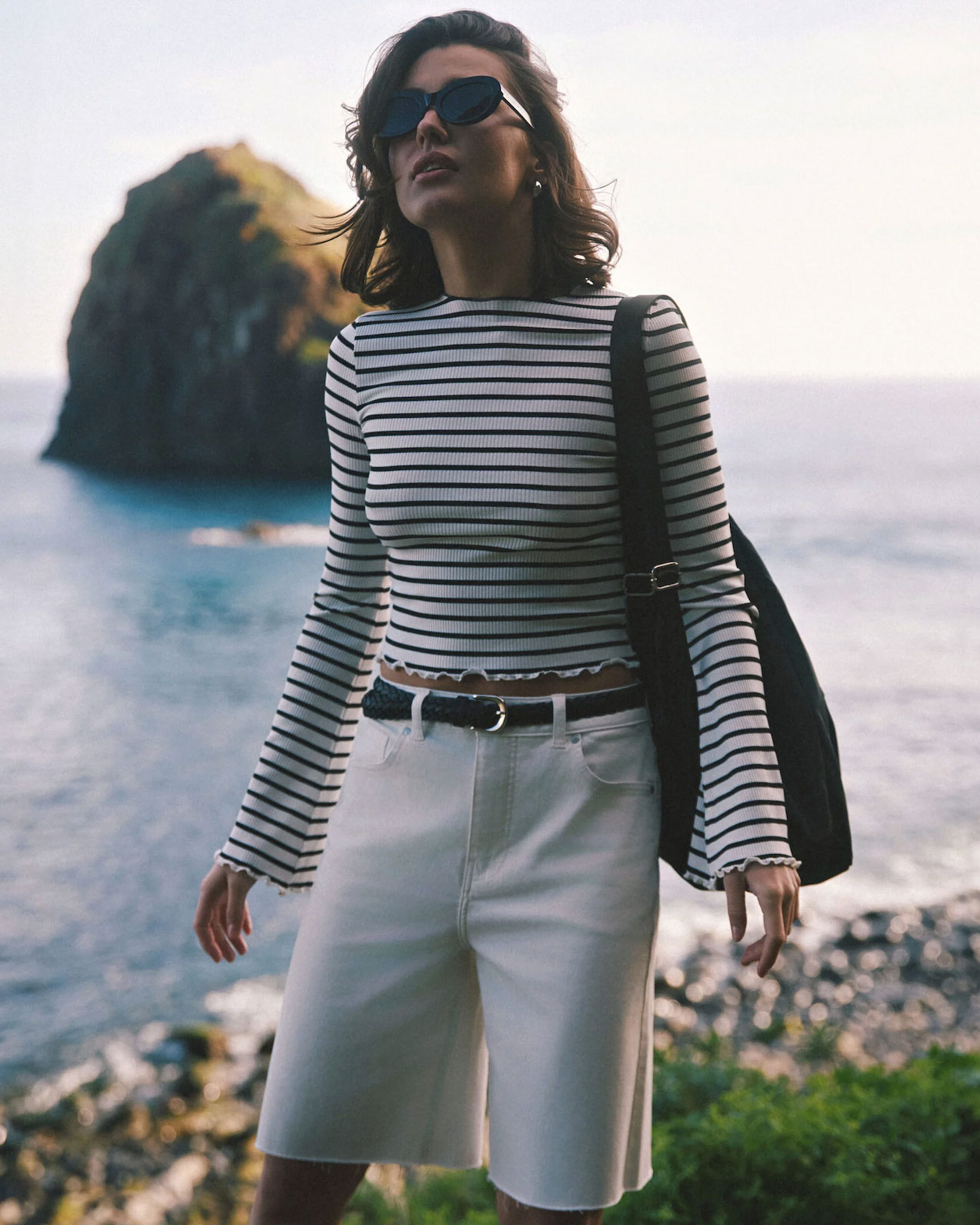Девушка стоит в полосатом лонгсливе Conte и белых шортах Conte на фоне океана. 