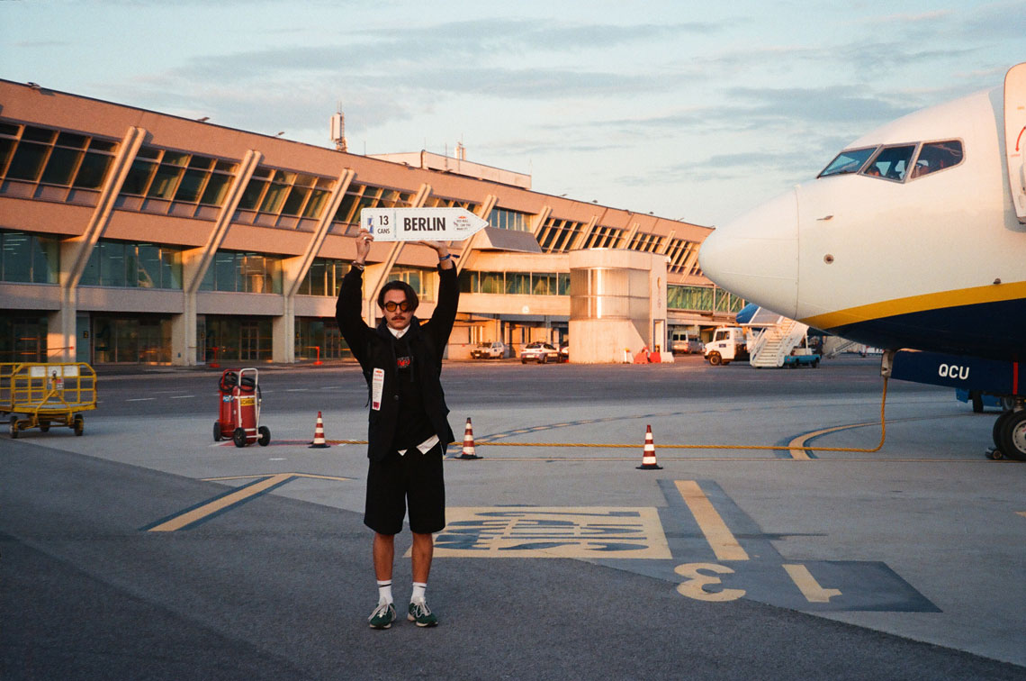 На фотографии изображен участник соревнования "Red Bull Can You Make It?" в аэропорту. Он стоит перед самолетом и держит табличку с надписью "Berlin" и числом "13". Парень одет в черный пиджак, белую рубашку и галстук.