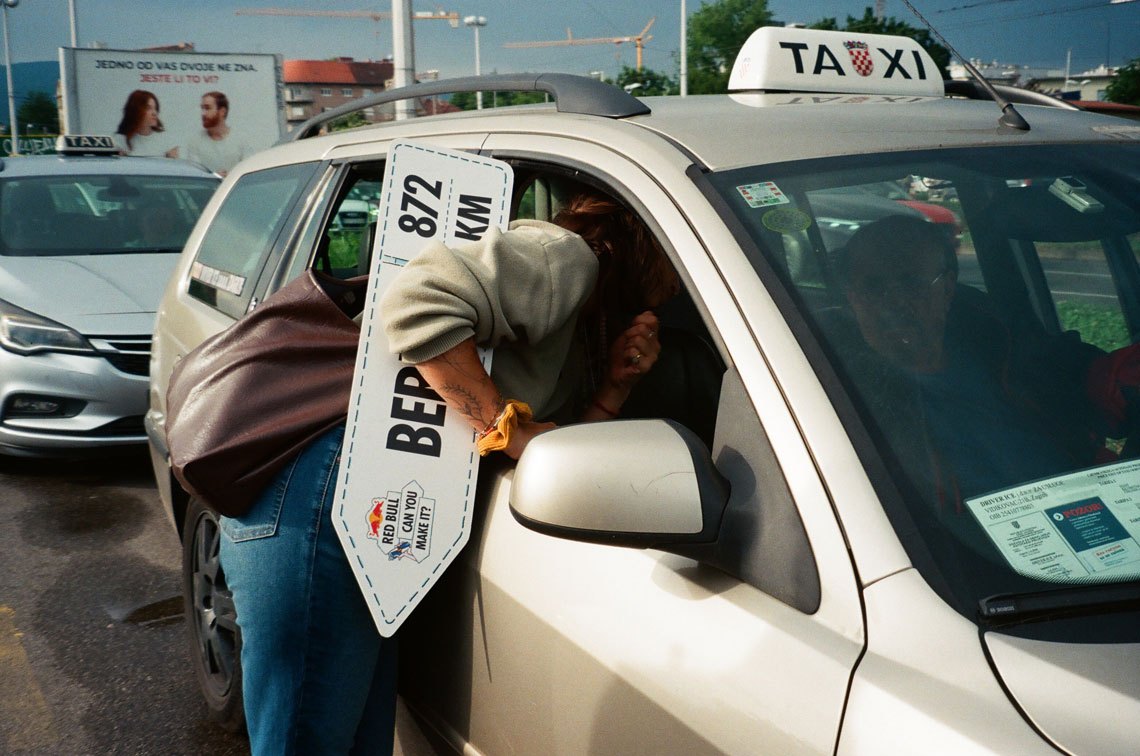 На фото парень что-то спрашивает у водителя такси. В руках он держит табличку с надписью "872 KM BERLIN" и логотипом "Red Bull Can You Make It?". Все происходит на парковке, рядом с другими автомобилями. 