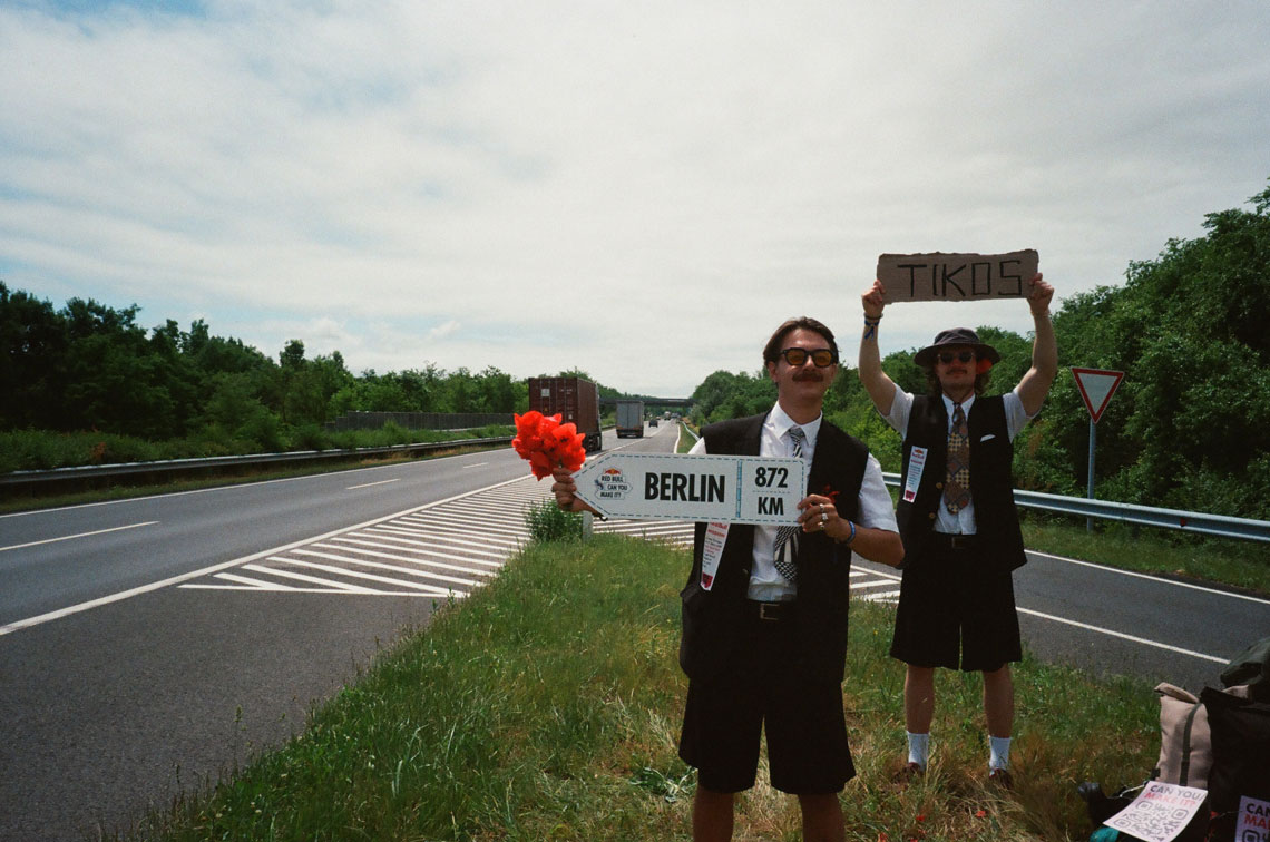 На фото двое парней стоят на обочине дороги с табличками "BERLIN 872 KM" и "TIKOS". Один держит букет искусственных цветов. Они одеты в черные жилеты, белые рубашки и галстуки. На заднем плане видно шоссе с проезжающими грузовиками. Они – участники проекта "Red Bull Can You Make It?"
