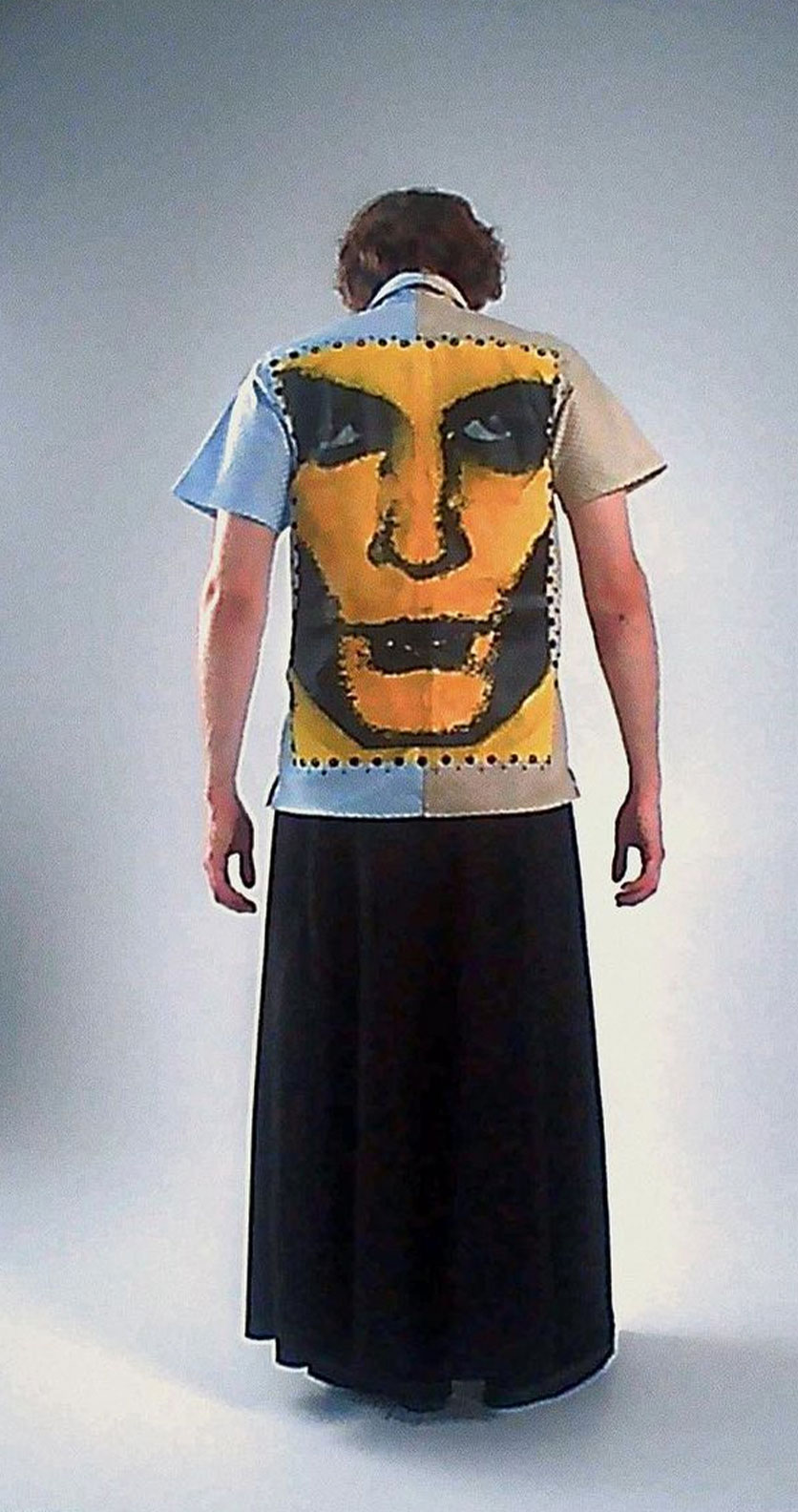 На фотографии изображен парень в длинной юбке и футболке с принтом. На футболке изображено лицо человека. Он стоит перед белой стеной.