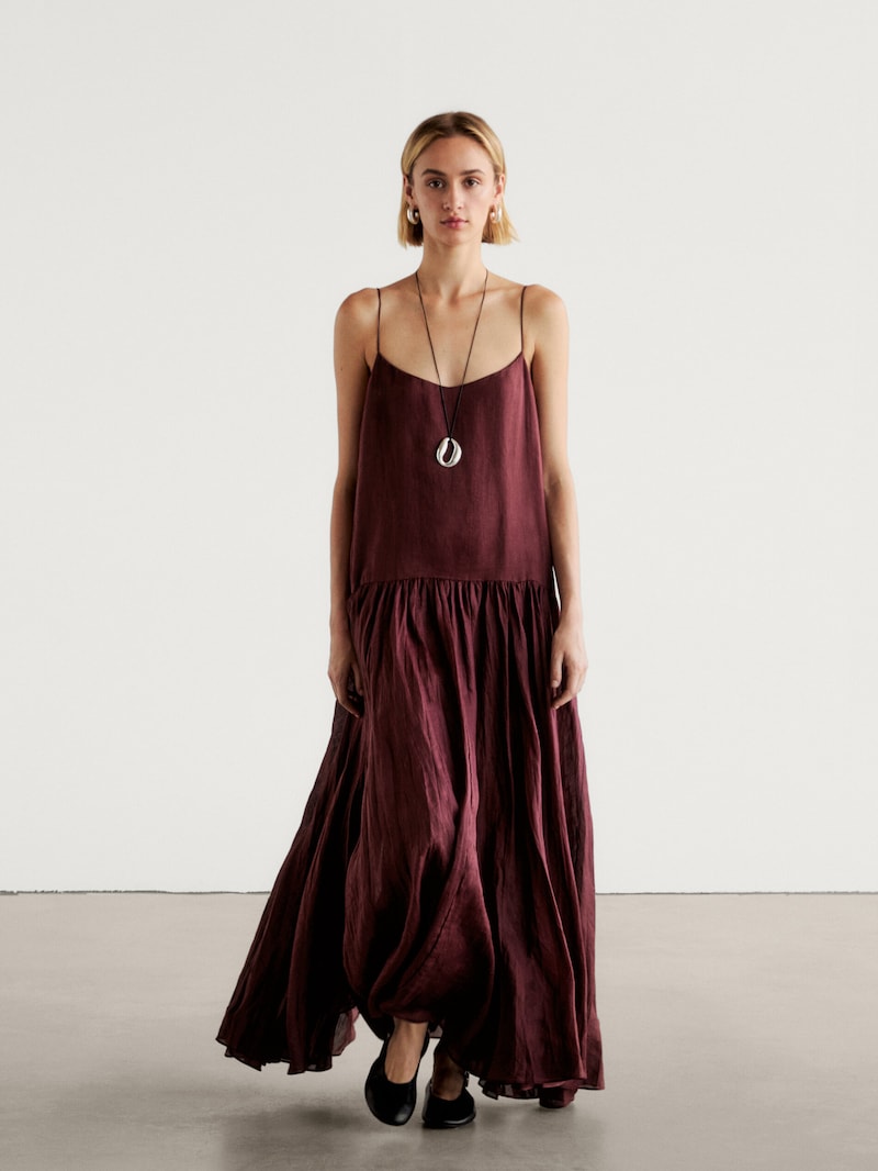Девушка в платье Massimo Dutti бордового цвета с тонкими бретелями и свободным силуэтом. Платье выполнено из легкой ткани с эффектом мятого шелка, что придает ему легкость и воздушность.